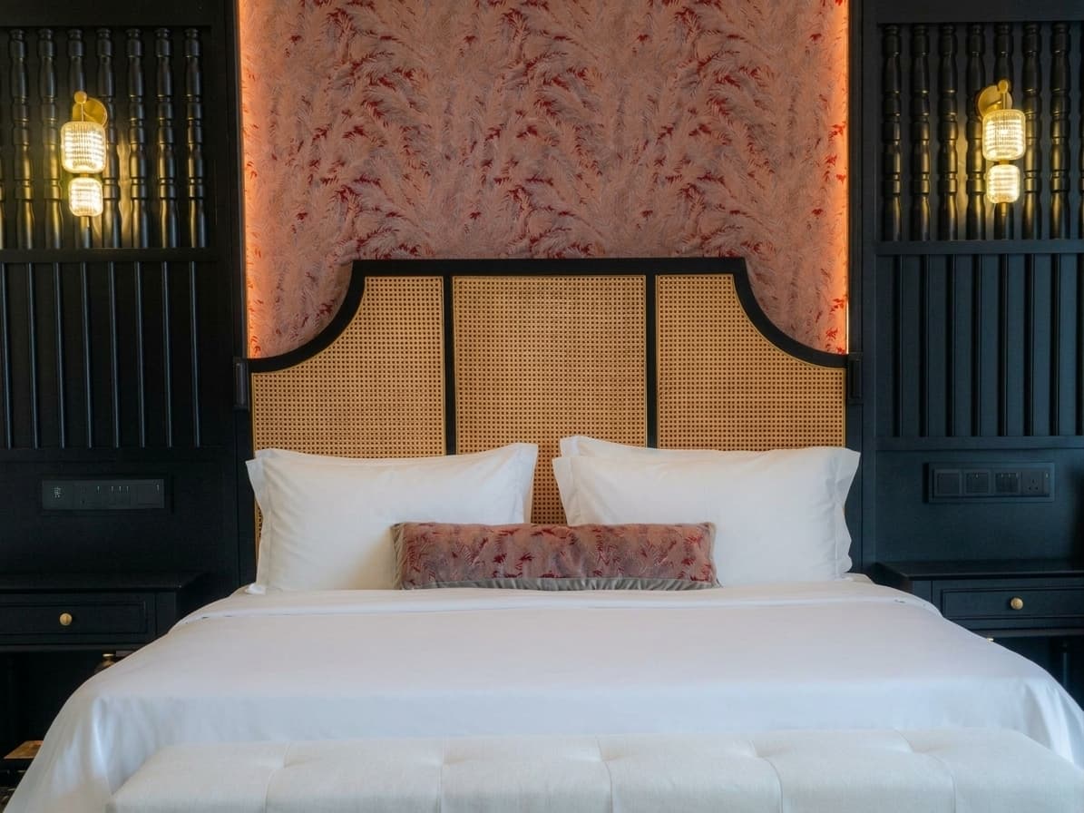 Photograph of a Deluxe Room: a caned arch headboard between black joinery, brass sconces and white linen.